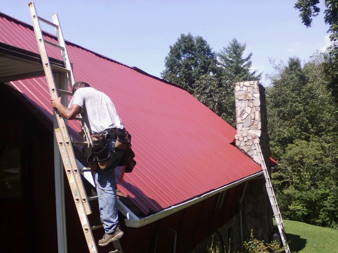 Roofer on a ladder beside a red metal roof and stone chimney — Soffit & Fascia Repair by Roofing Brothers in Parrish